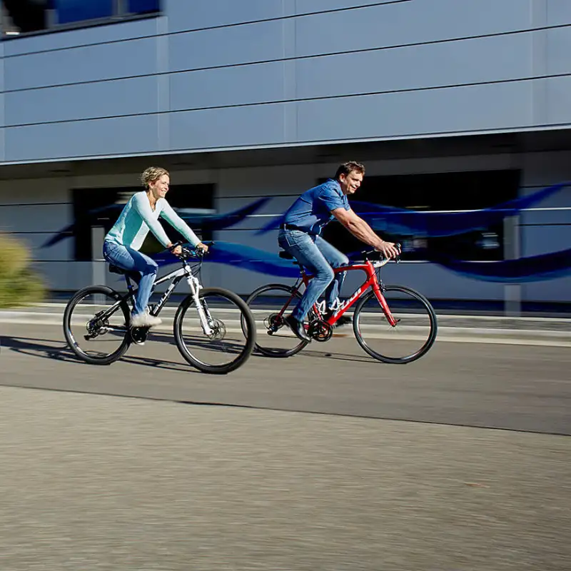 Zwei Menschen, die auf zwei Stadträdern vor einem Büro-Gebäude vorbeifahren Zwei Menschen, die auf zwei Stadträdern vor einem Büro-Gebäude vorbeifahren