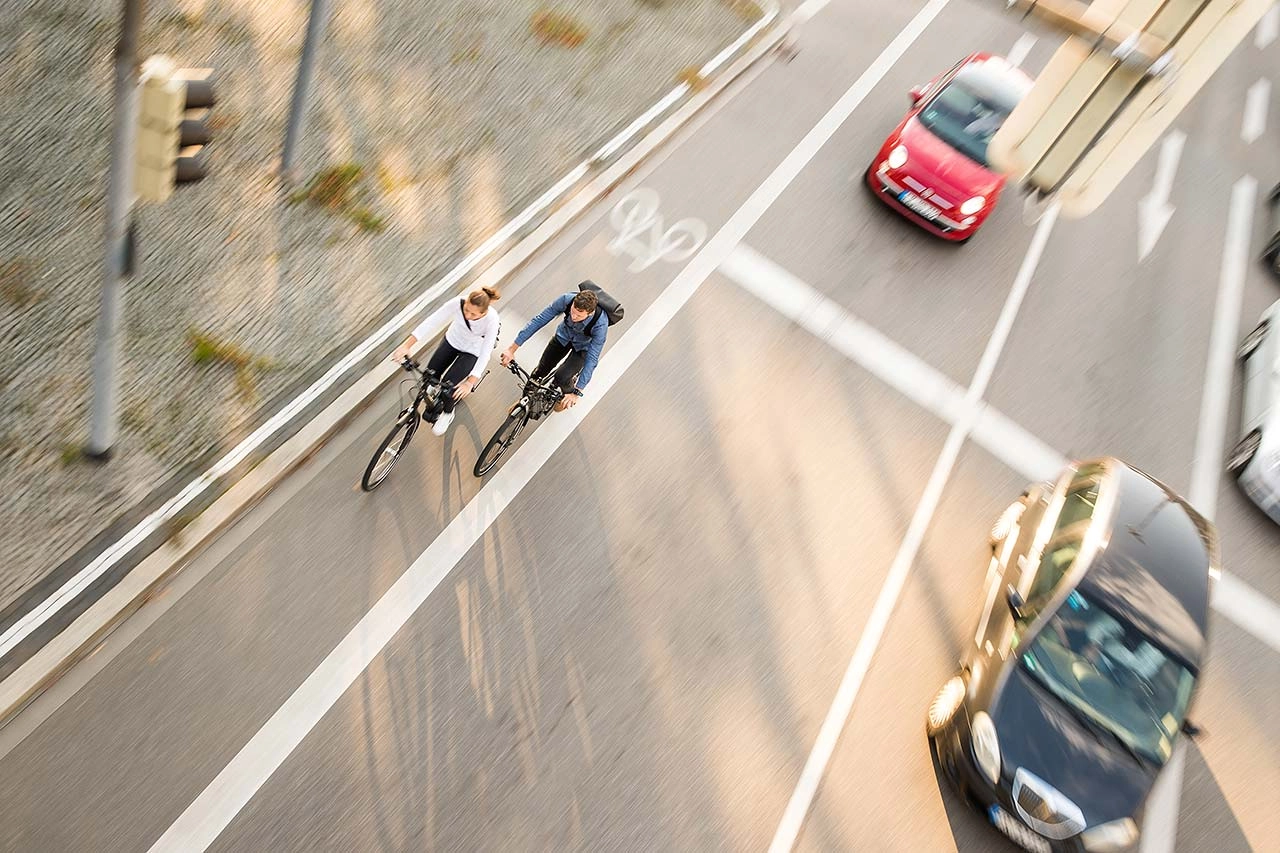 Zwei Fahrradfahrer:innen im Straßenverkehr mit Autos, von schräg vorne/oben fotografiert